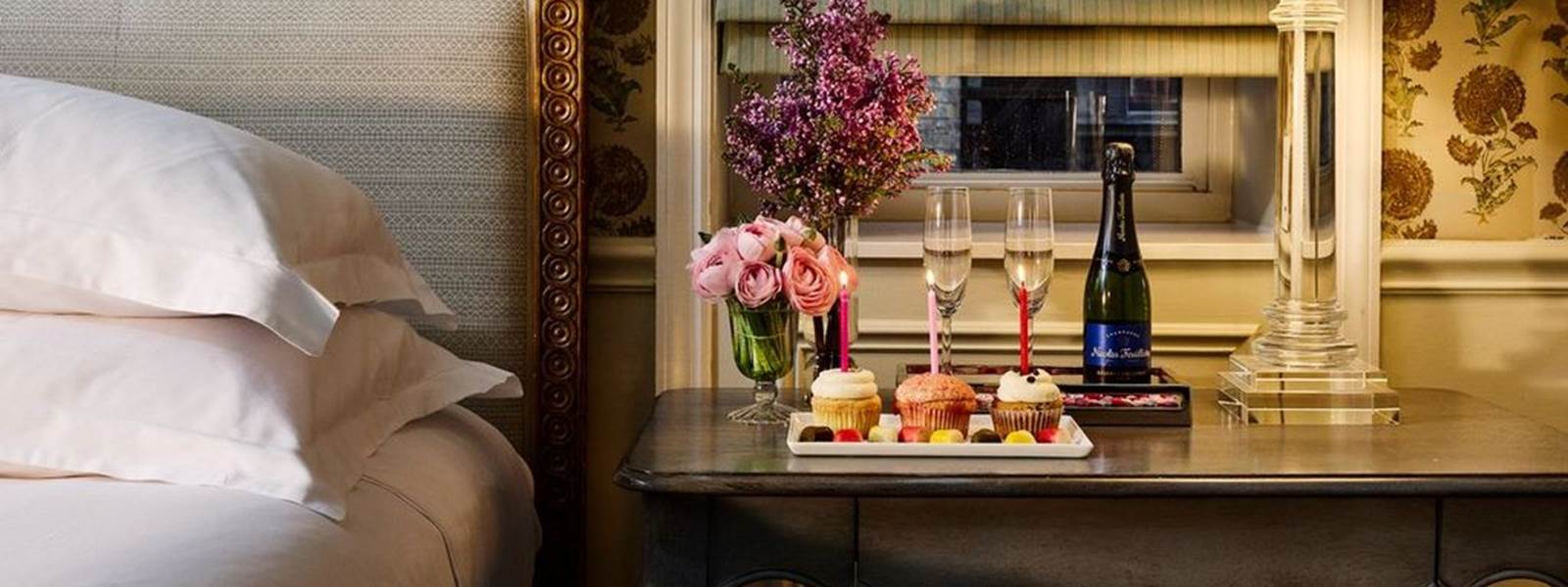 a bedside table with champagne, cupcakes, and flowers