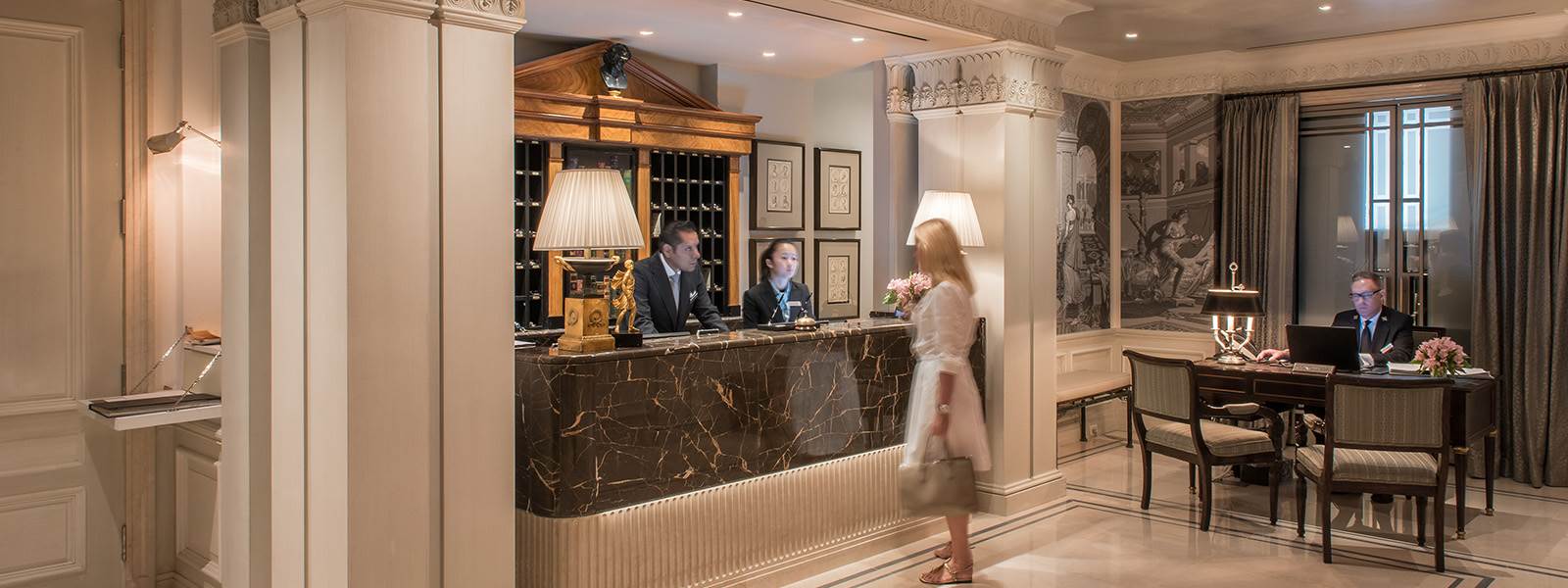 A woman approaches the marble reception desk in a luxurious lobby with a man and woman behind the counter and another man sitting at a desk.